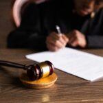 An individual blurred in the background writes by a gavel on a table, suggesting a legal setting for a debtor examination process.