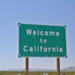 The image features a road sign with the message "Welcome to California" prominently displayed in white capital letters against a green background. The sign shows signs of wear and tear, set against a clear blue sky with a mountain range faintly visible in the distance.