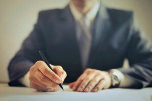 A focused close-up of a man's hands holding a pen, signing documents at a desk.