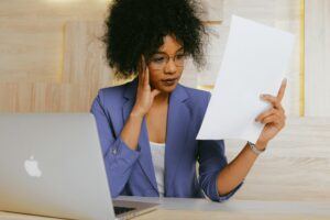 A woman examines a document thoughtfully while working on her laptop in a modern office.