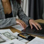 A woman works at a computer, with a calculator to her side, making adjustments that pertain to the money and documents also beside her.