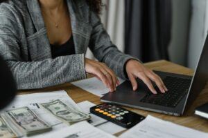 A woman works at a computer, with a calculator to her side, making adjustments that pertain to the money and documents also beside her.
