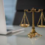 Brass balance scale placed beside a laptop on a glass desk, symbolizing justice, legal practice, and debt collection enforcement in a professional setting.