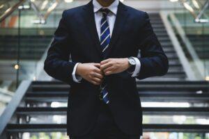 Business professional in a dark suit buttoning jacket, standing confidently in front of a staircase — representing expertise in judgment enforcement and debt collection.
