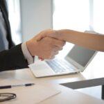 Close-up of a professional handshake across a desk with a laptop, pen, and glasses, representing agreement or partnership in a legal or financial setting.