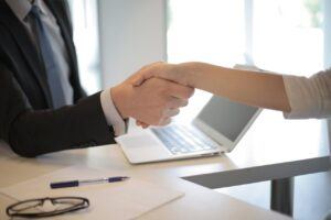 Close-up of a professional handshake across a desk with a laptop, pen, and glasses, representing agreement or partnership in a legal or financial setting.