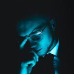 Man in glasses and a suit bathed in moody blue light, appearing deep in thought or stress, symbolizing financial pressure or legal uncertainty.
