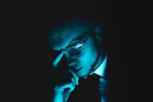 Man in glasses and a suit bathed in moody blue light, appearing deep in thought or stress, symbolizing financial pressure or legal uncertainty.