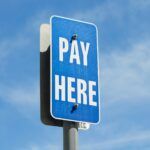 Close-up of a blue “Pay Here” parking sign mounted on a pole, set against a sunny sky with scattered clouds.