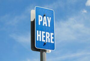 Close-up of a blue “Pay Here” parking sign mounted on a pole, set against a sunny sky with scattered clouds.